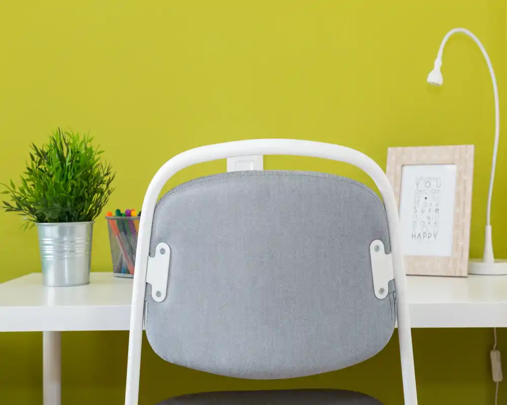 Close-up of a desk and chair against a bright green wall, styled with a small potted plant, pencil holder, framed print and a flexible desk lamp — a neat, well-lit workspace corner.