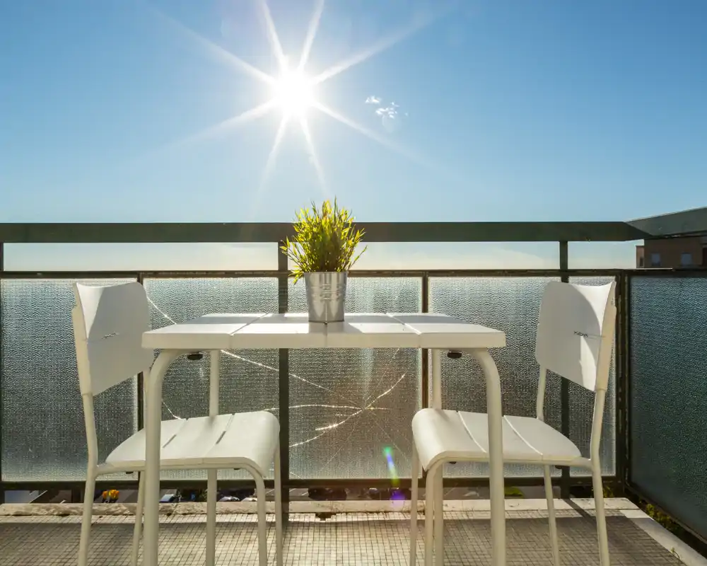 Sunny private balcony with a small white table and two chairs facing a glass railing — ideal for morning coffee with clear skies and bright light.