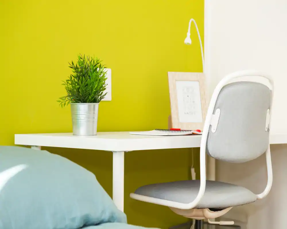 Bright, modern bedroom corner with a small white desk, grey chair and a hint of the bed in the foreground; tidy workspace with a potted plant and framed print against a lively green wall.
