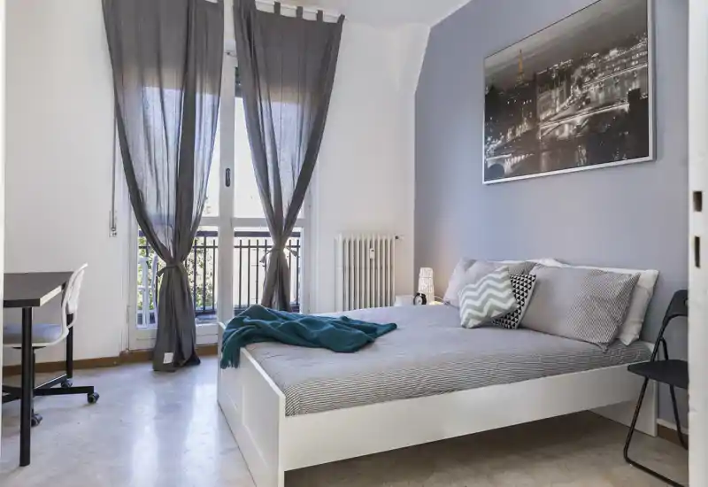 Bright, modern bedroom with a white platform bed, patterned cushions and a soft throw; large window with sheer curtains brings natural light to the room.