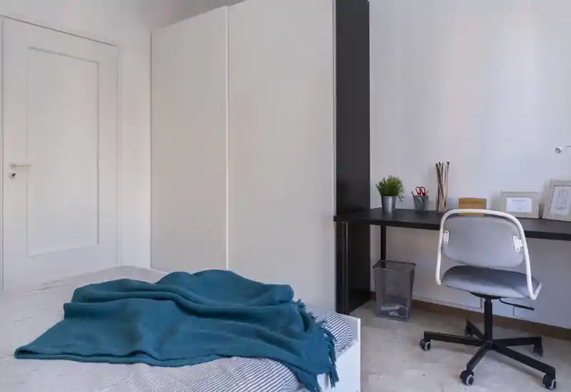Bright, minimal bedroom with a visible bed covered by a teal throw, a large white wardrobe and a simple desk with chair — a clean, modern space ideal for short-term rentals.
