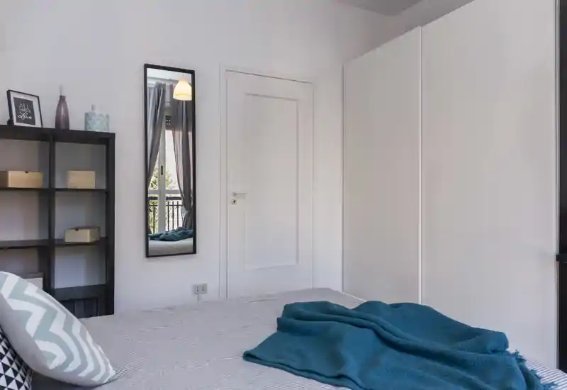 Bright, modern bedroom with a neatly made bed, teal throw and patterned cushions; simple shelving and a tall mirror create a functional, airy space.