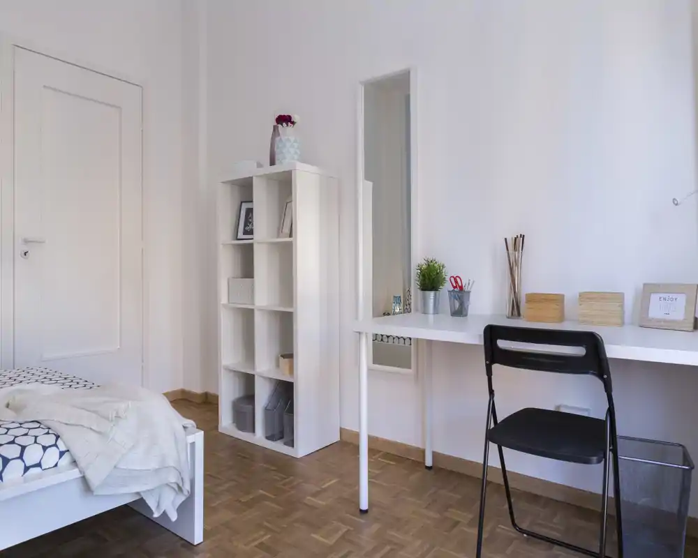 Chambre claire et minimaliste avec un lit blanc, des étagères ordonnées et un petit bureau — idéale pour une personne ou un étudiant. La lumière naturelle douce et la décoration épurée apportent une atmosphère calme et accueillante.