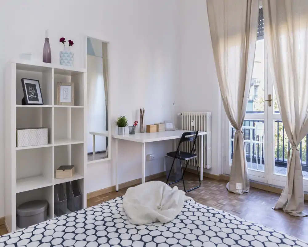 Chambre moderne et claire avec une couette à motifs, un bureau ordonné près du balcon et des étagères ouvertes pour le rangement — lumineuse et aérée grâce à la lumière naturelle.