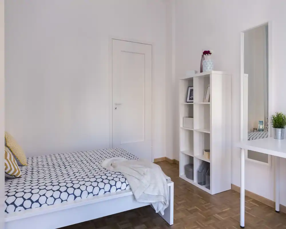 Chambre lumineuse et minimaliste avec un lit double à motif, sol en bois clair et une étagère blanche; style moderne et épuré, idéal pour les annonces.