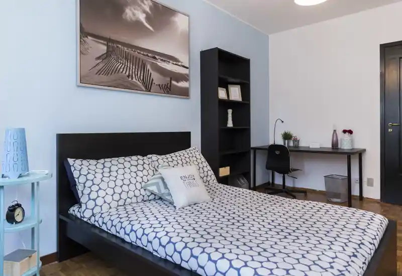 Bright, stylish bedroom with a double bed dressed in patterned linens, bedside table and a small workspace with desk and shelving — cozy and well organized for guests or long stays.