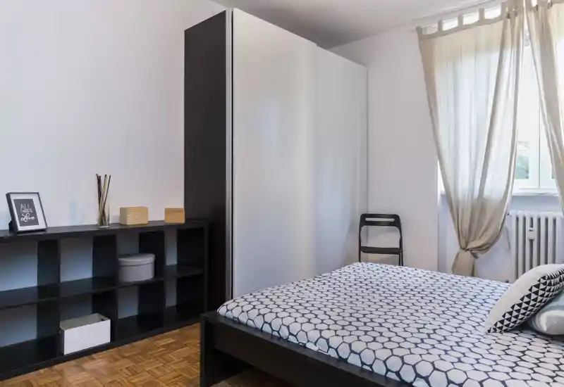 Bright, simply styled bedroom with a patterned double bed, wardrobe and shelving. Natural light from the window and neutral decor create a calm atmosphere ideal for listings.