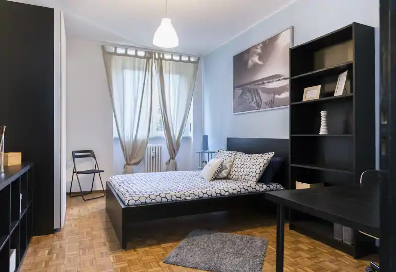 Bright, modern bedroom with a double bed, patterned bedding, shelving and natural light from a window — calm and well-styled for rental listings.