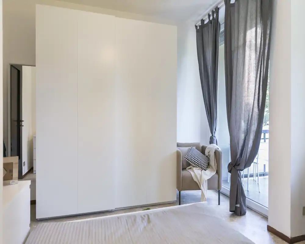 Bright, modern bedroom with a large white wardrobe, a portion of the bed in the foreground and a cozy armchair by a floor-to-ceiling window with sheer curtains.