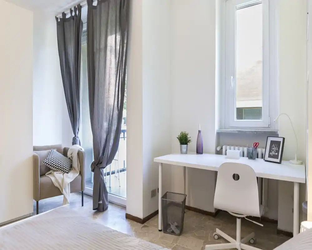 Bright, modern bedroom with a neat white desk by the window, a comfortable bed in the foreground and a cozy armchair by the glass door leading to a small balcony.