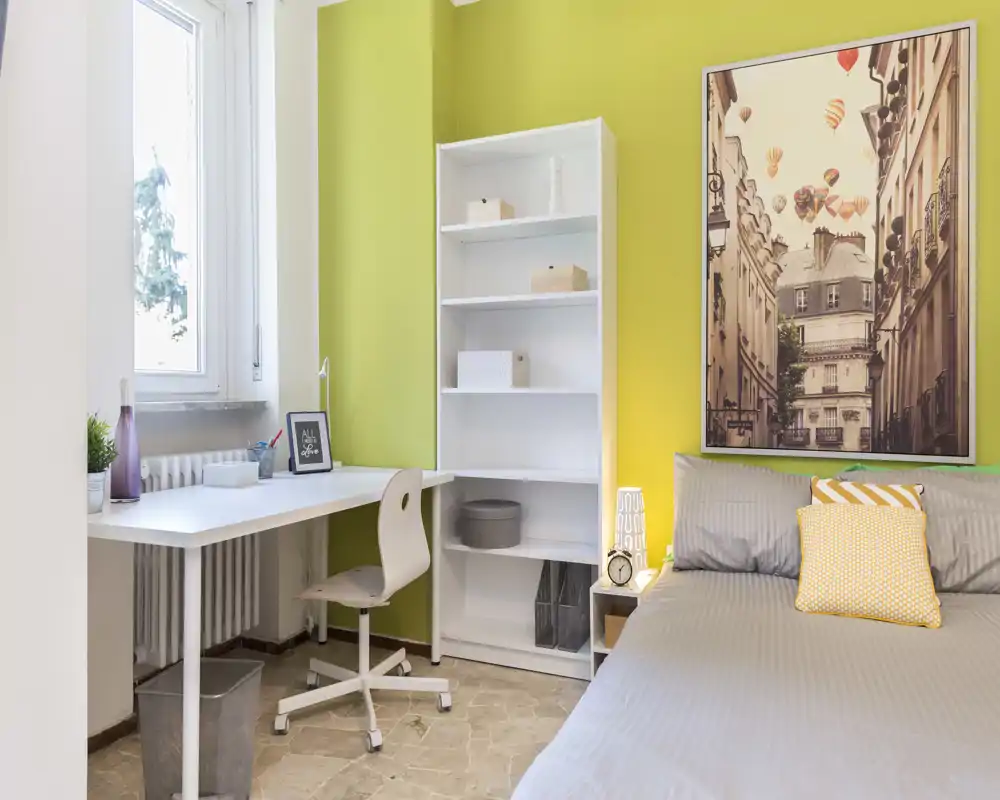 Bright, modern bedroom with a neatly made bed, bedside lamp and a workspace by the window — ideal for both rest and study.