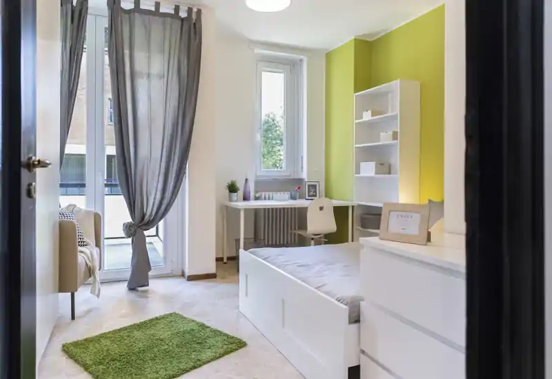 Bright, modern bedroom with a white bed frame, desk by the window, shelving and a green accent wall creating a fresh, welcoming atmosphere.