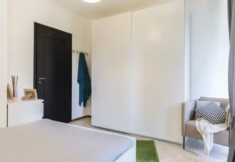Bright modern bedroom with a large white wardrobe, a neatly made bed visible in the foreground and a cozy armchair by the window.