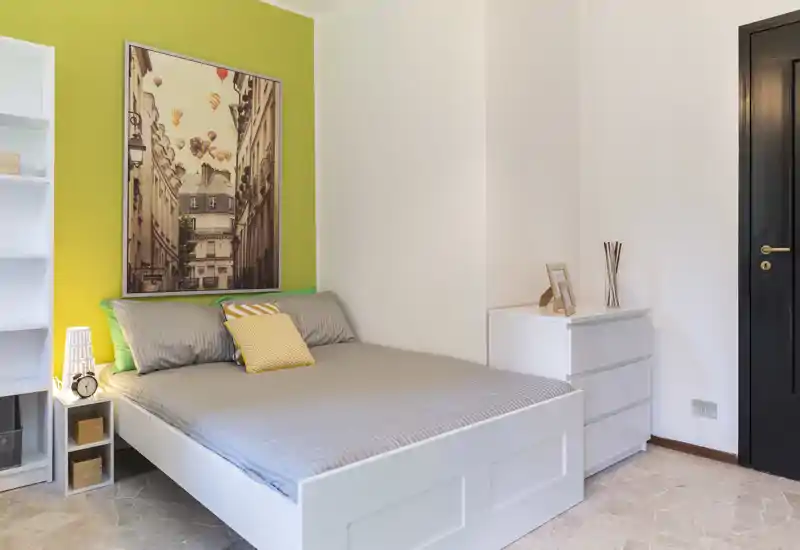 Bright, modern bedroom with a full white bed, soft gray bedding and colorful accent pillows against a lime-green feature wall; neat storage and a dresser complete the space.