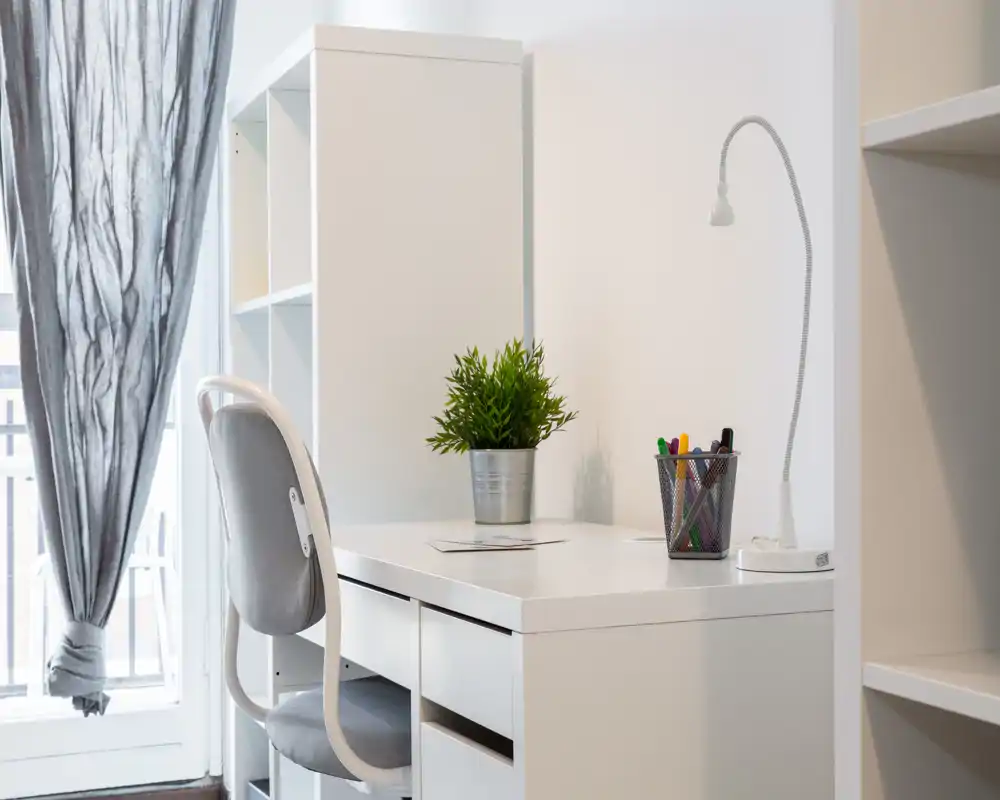 Rincón de estudio luminoso y minimalista con escritorio blanco, estantería, silla y una pequeña planta en maceta — ideal para trabajar o estudiar.