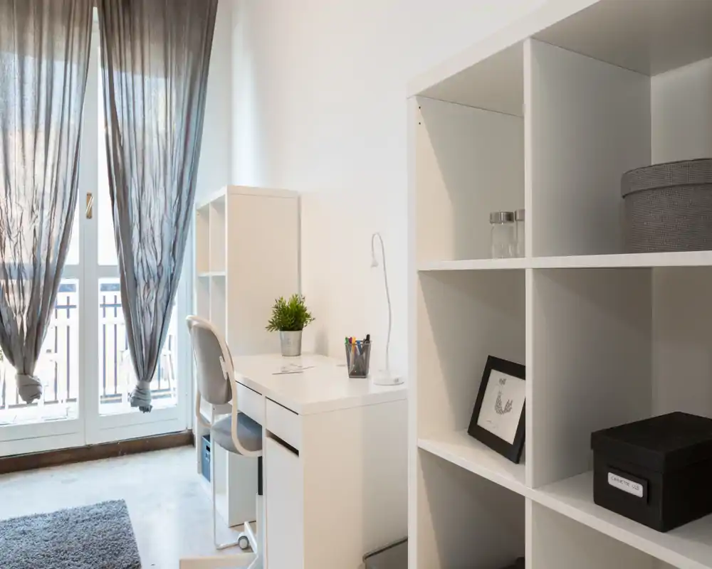 Zona de estar luminosa y moderna con escritorio ordenado y estantería junto a una gran ventana — ideal para teletrabajo o relajarse en un espacio minimalista y diáfano.