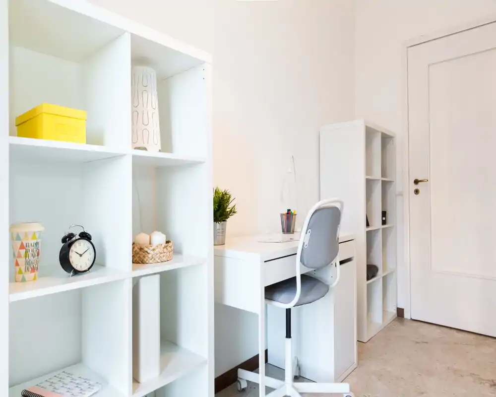 Rincón de estudio ordenado con escritorio blanco, silla giratoria y estanterías abiertas; luminoso, minimalista y bien organizado — ideal para teletrabajo o estudio.