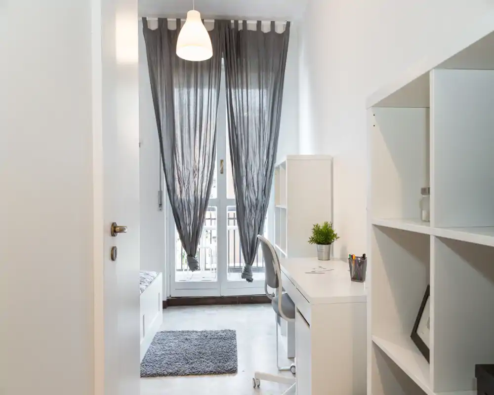 Dormitorio luminoso y minimalista con rincón de estudio: escritorio y silla blancos, estanterías, alfombra suave y gran ventana con cortinas oscuras semitranslúcidas que dejan pasar la luz natural.