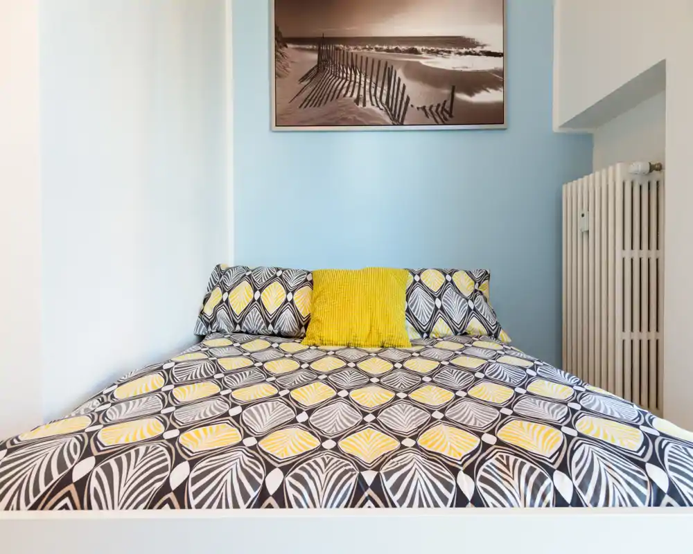 Dormitorio luminoso y bien presentado con una amplia cama estampada, cojín amarillo y pared azul suave — perfecto para anuncios que destaquen confort y estilo moderno.