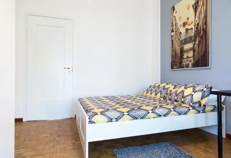 Dormitorio luminoso y sencillo con cama doble en estructura blanca, ropa de cama estampada, suelo de parquet y cuadro en la pared — ideal para alquileres de corta estancia.