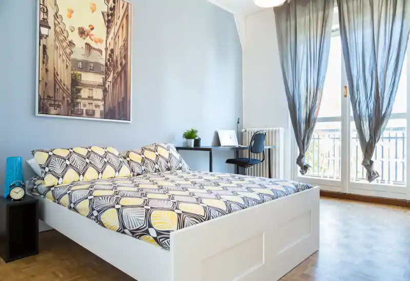 Dormitorio luminoso y con estilo, con una cama doble de marco blanco, ropa de cama estampada y una gran ventana que aporta luz natural. Un pequeño escritorio junto a la ventana es perfecto para trabajar desde casa.