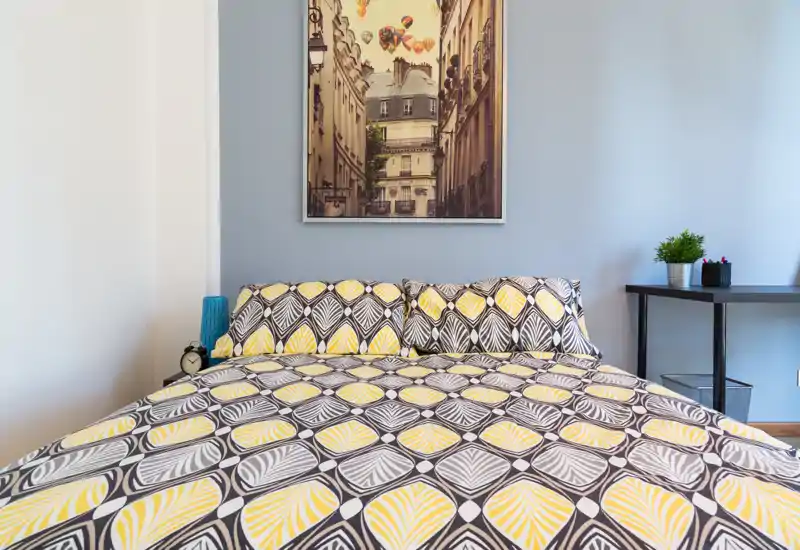 Dormitorio luminoso y decorado con edredón estampado y cojines, cuadro sobre la cama y una mesita con planta y accesorios.