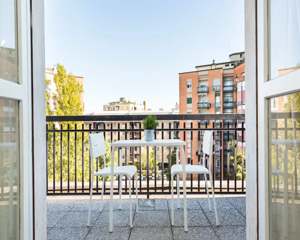 Balcone soleggiato con piccolo tavolo e due sedie che si affacciano sugli edifici della città — ideale per un caffè al mattino o per rilassarsi all'aperto.