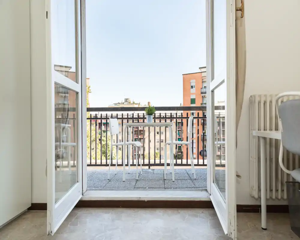 Balcone soleggiato visto attraverso porte finestre aperte, con un piccolo tavolo e sedie e vista sugli edifici vicini—ideale per il caffè del mattino.