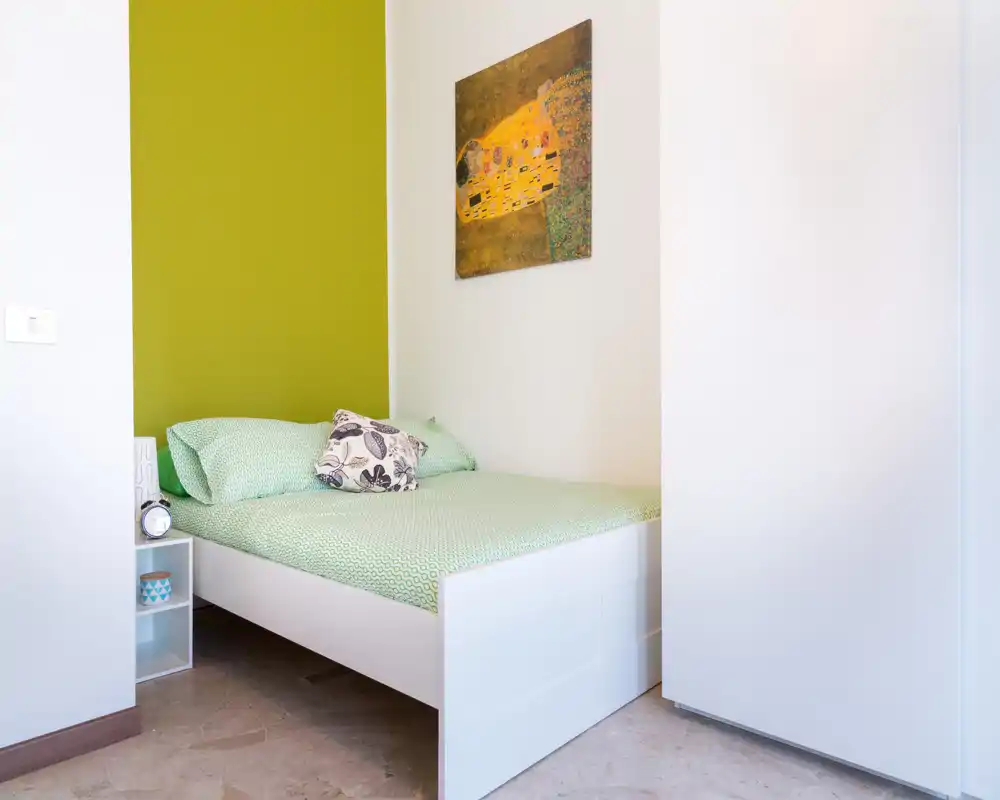 Camera da letto compatta e luminosa con un letto singolo ben rifatto contro una parete verde, comodino a giorno e quadro decorativo — perfetta per un soggiorno accogliente.