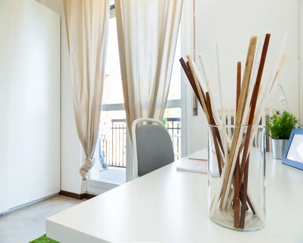 Angolo living luminoso e moderno con scrivania bianca, vaso decorativo con bastoncini, sedia e tende leggere che fanno filtrare la luce naturale — stile minimal e arioso, adatto a lavoro o lettura.