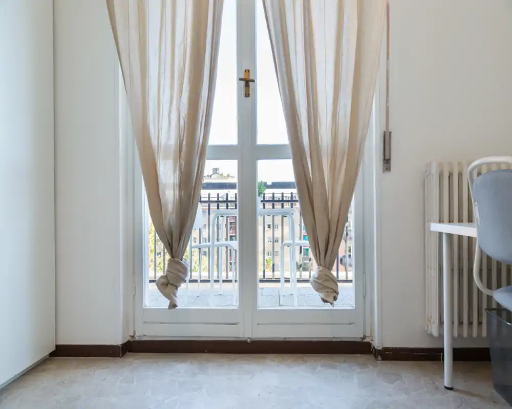 Vista interna luminosa e minimale focalizzata su una porta finestra che dà su un piccolo balcone; tende leggere annodate lasciano filtrare la luce naturale.