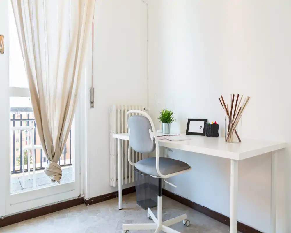 Spazio di lavoro luminoso e minimale vicino al balcone: scrivania bianca, sedia girevole, pianta e accessori ordinati per un angolo studio rilassante.