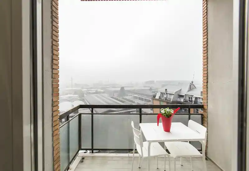 Piccolo balcone con tavolo e sedie bianchi e un vaso rosso a forma di annaffiatoio; vista sulla città. Perfetto per un caffè mattutino o un'area esterna compatta.