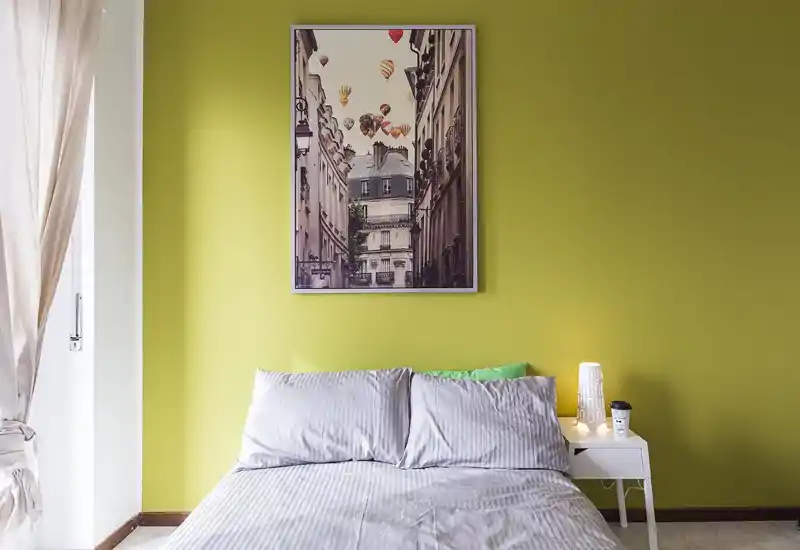 Camera da letto moderna e luminosa con letto matrimoniale ben disposto davanti a una parete verde vivace e un quadro appeso; comodino con lampada e tazza per praticità.