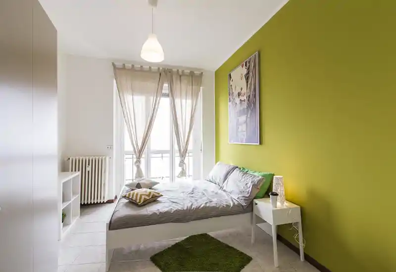 Camera da letto luminosa e moderna con letto matrimoniale comodo, parete accentuata verde e ampie finestre con tende leggere. Arredi minimali con comodino, lampada e cuscini decorativi per un'atmosfera accogliente.