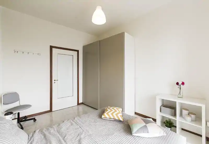 Camera da letto luminosa e minimal con un grande letto in primo piano, armadio neutro, mensola semplice e cuscini decorativi per un'atmosfera accogliente.