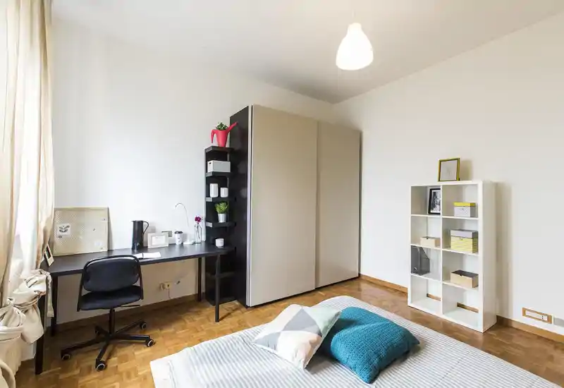 Dormitorio luminoso y minimalista con cama baja, ropa de cama a rayas y cojines coloridos, escritorio y armario que ofrecen almacenamiento y zona de trabajo.