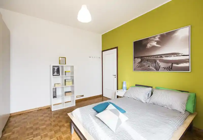 Dormitorio moderno y luminoso con cama doble frente a una pared verde, ropa de cama a rayas, mesita con lámpara y un cuadro marino encima; suelo de parquet y estantería sencilla completan el ambiente.