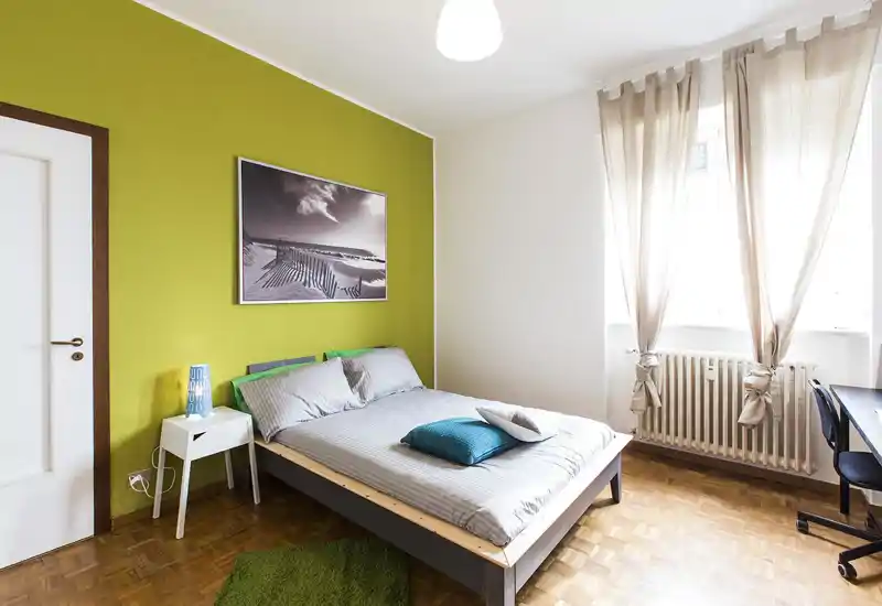 Dormitorio moderno y luminoso con cama doble baja de madera vestida con ropa de cama gris, cojines decorativos y pared verde; amplia ventana con cortinas ligeras que deja entrar mucha luz natural.