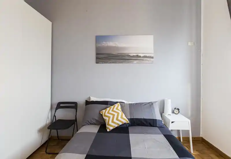 Dormitorio luminoso y minimalista con cama doble vestida en ropa de cama gris de cuadros, cojín decorativo en zigzag, mesita con lámpara y despertador, y un cuadro del mar sobre la cabecera.