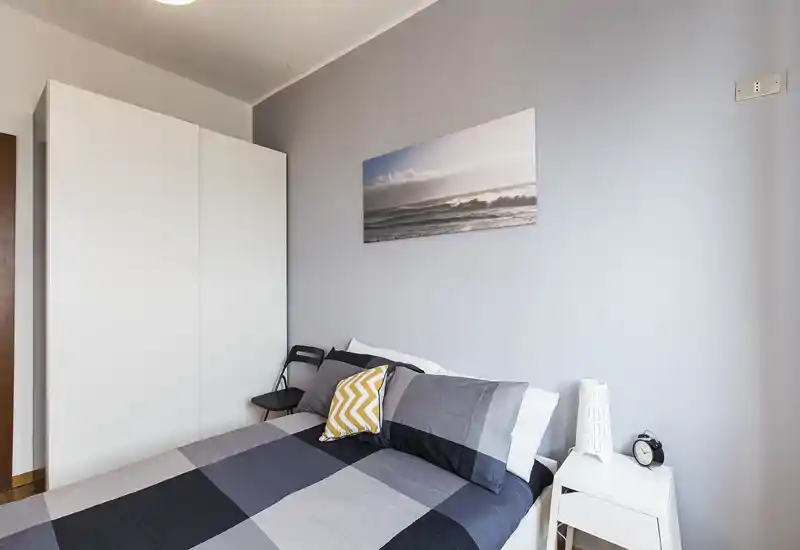 Dormitorio moderno y ordenado con cama doble con ropa de cama gris, cojín decorativo y armario blanco. La luz natural suave y el cuadro costero refuerzan una sensación tranquila y minimalista, perfecto para descansar.