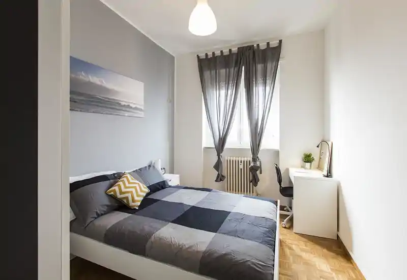 Dormitorio luminoso y bien presentado con una cama doble grande con edredón gris a cuadros, un cojín decorativo amarillo y un pequeño escritorio junto a la ventana.