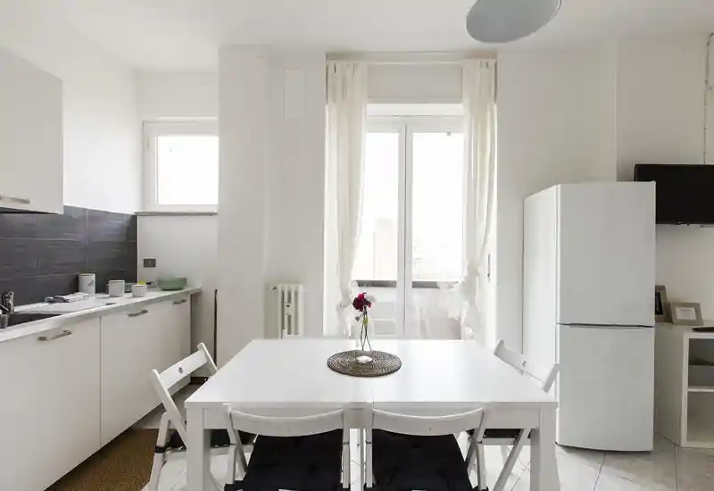 Cocina moderna y luminosa con mesa blanca, cuatro sillas y frigorífico; una ventana/puerta grande aporta mucha luz natural y un centro de mesa sencillo añade calidez.
