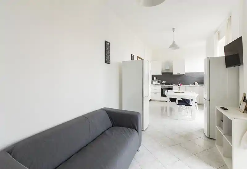 Sala de estar abierta y luminosa con sofá gris y mesa de comedor pequeña que conecta con una cocina blanca — estilo minimalista y limpio, ideal para alquileres temporales.