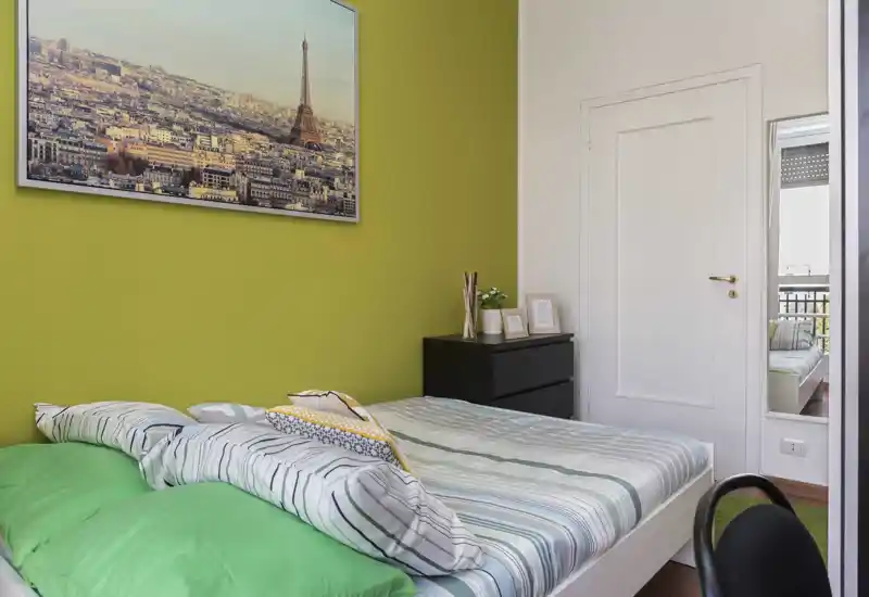 Dormitorio luminoso y bien preparado con cama doble frente a una pared verde, cojines decorativos y una mesita negra; la luz natural y un cuadro enmarcado crean un ambiente acogedor.