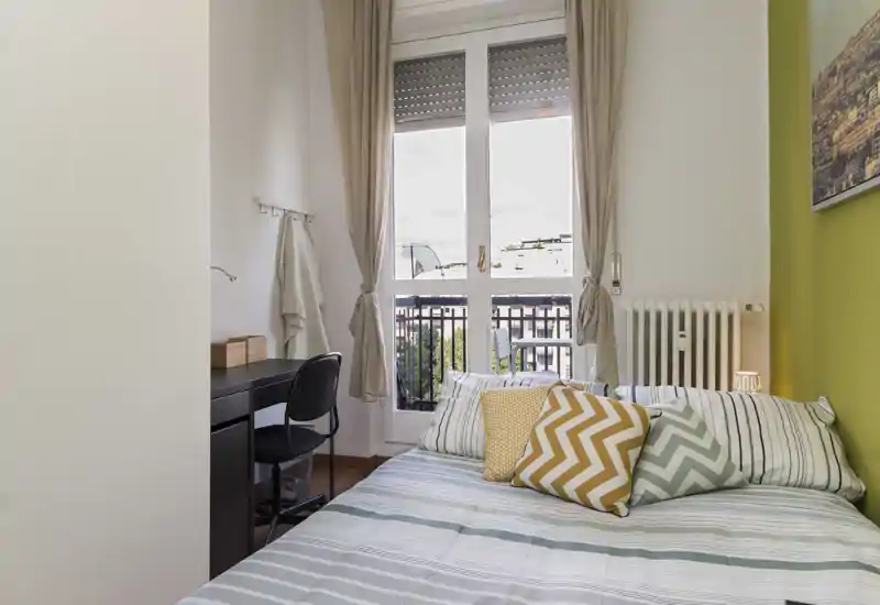 Dormitorio luminoso y ordenado con cama doble, cojines decorativos y un escritorio junto a la ventana con salida al balcón.
