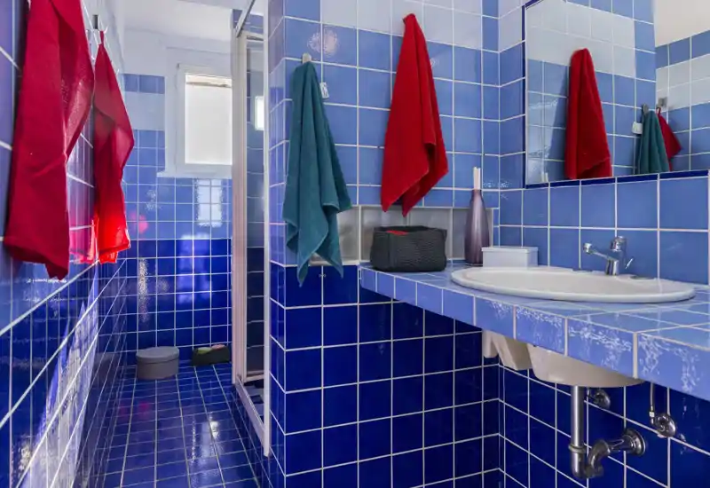 Bright, modern bathroom with blue ceramic tiles, a sink with mirror, and a shower cubicle; colorful towels add a vibrant touch.
