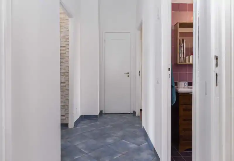 Bright, narrow hallway with blue tiled floor and white walls leading to multiple doors; clean, minimalist entrance corridor.