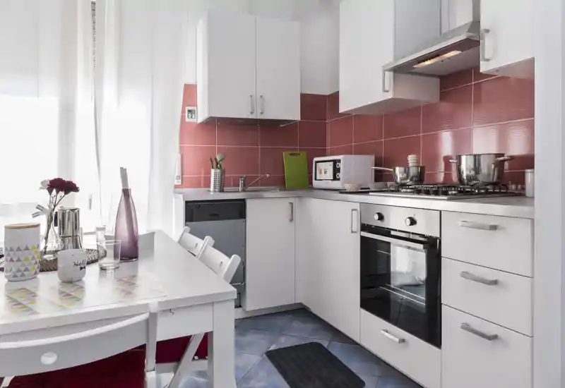Bright, modern kitchenette with white cabinets, red tile backsplash, built-in oven and a small dining table set by the window.