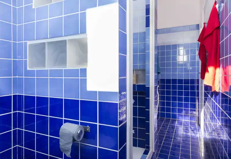 Bright blue-tiled bathroom with a shower area, towel hooks and toilet paper holder visible; glossy ceramic tiles create a reflective, clean look.